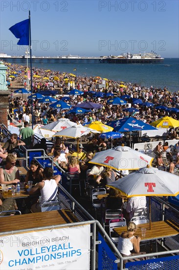 ENGLAND, East Sussex, Brighton, People sitting under sun shade umbrellas at tables on the promenade outside bars and restaurants with Brighton Pier and people on the shingle pebble beach beyond.