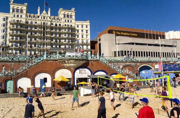 ENGLAND, East Sussex, Brighton, Young people playing beach volleyball on sand on the seafront with the De Vere Grand Hotel and The Brighton Centre beyond.