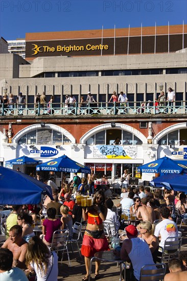 ENGLAND, East Sussex, Brighton, People sitting under sun shade umbrellas at tables on the promenade outside the Gemini Beach Bar with The Brighton Centre beyond.