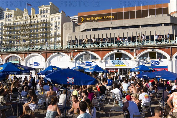 ENGLAND, East Sussex, Brighton, People sitting under sun shade umbrellas at tables on the promenade outside the Gemini Beach Bar with the De Vere Grand Hotel and The Brighton Centre beyond.