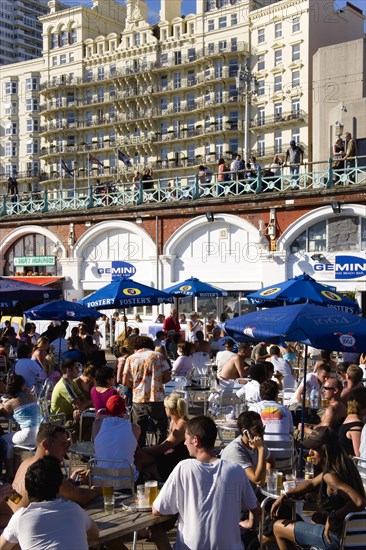 ENGLAND, East Sussex, Brighton, People sitting under sun shade umbrellas at tables on the promenade outside the Gemini Beach Bar with the De Vere Grand Hotel beyond.