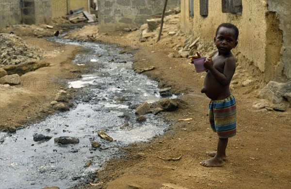 Ghana, Dixcove, Child holding drinking cup standing beside open sewer through street centre.