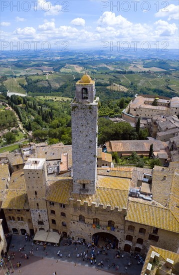 ITALY, Tuscany, San Gimignano, View of the Palazzo Vecchio del Podesta of 1239 with its medieval tower with tourists in the Piazza del Duomo with rooftops and Tuscan farmland on the slopes of the hill town.