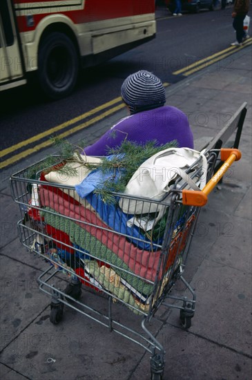 England, East Sussex, Brighton, Homeless lady sitting on a pavement bench beside a road with a bus passing and her possessions in a supermarket trolley beside her.