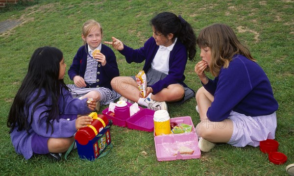 England, East Sussex, Brighton, Four young schoolgirls sitting on the grass in the school playing fields having their home packed lunch meals from their lunchboxes.