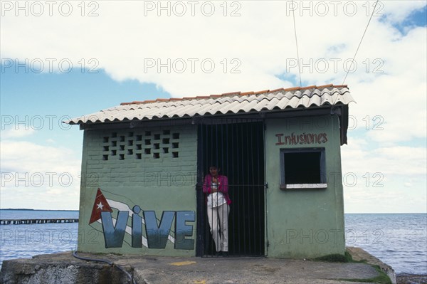 CUBA, West Coast, Vinales,Woman standing in doorway of drinks store building with Vive painted on exterior. The ocean behind.