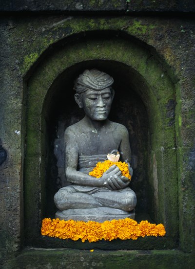 Indonesia, Bali, Ubud, Stone statue of male figure in niche of wall sitting in lotus position holding and surrounded by yellow frangipani flowers.