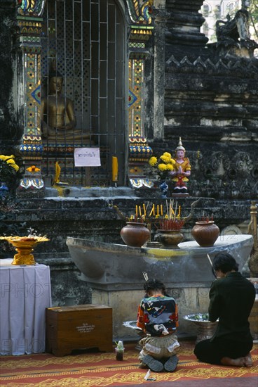 Thailand, North, Chiang Mai, Wat Chettawan Buddhist temple on Tha Phae Road with woman and young girl kneeling and holding incense sticks before a seated statue of Buddha in a niche at an outdoor shrine.