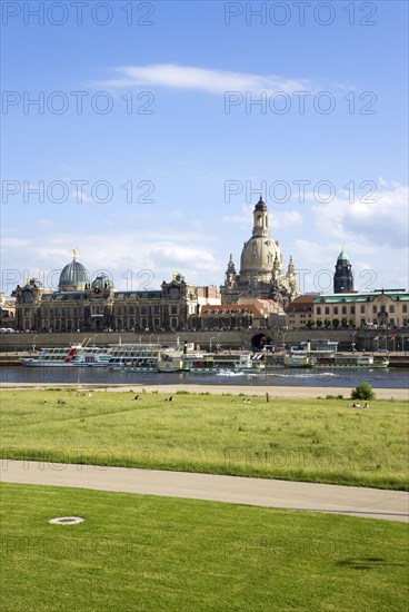 GERMANY, Saxony, Dresden, The city skyline with cruise boats moored on the River Elbe in front of the embankment buildings on the Bruhl Terrace busy with tourists of the Art Academy the restored Baroque Frauenkirche Church of Our Lady Dome with people sitting on the grass of the near river embankment.