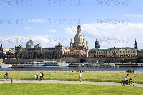 GERMANY, Saxony, Dresden, The city skyline with cruise boats moored on the River Elbe in front of the embankment buildings on the Bruhl Terrace busy with tourists of the Art Academy the restored Baroque Frauenkirche Church of Our Lady Dome with some people sitting on the grass or cycling or walking on the path of the near river embankment.