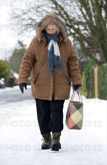 England, West Sussex, Chichester, Middles aged woman in warm clothing carrying shopping bag and walking carefully along icy pavement in winter snow.