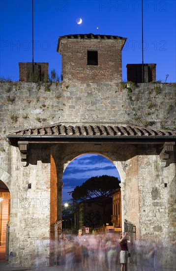 ITALY, Tuscany, Pisa, The Campo dei Miracoli or Field of Miracles.The west entrance gate at dusk with tourists passing through with a half crescent moon in the clear blue sky.