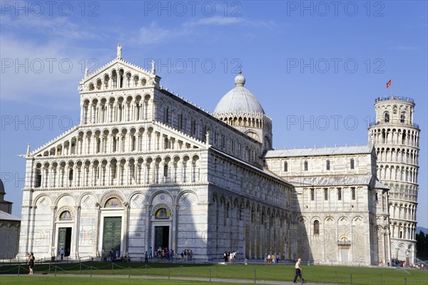 ITALY, Tuscany, Pisa, Campo dei Miracoli or Field of Miracles The Duomo Cathedral and Leaning Tower belltower or Torre Pendente under a blue sky with tourist walking at their base.