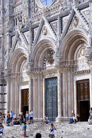 ITALY, Tuscany, Siena, Tourists on the steps of the pink black and white marble facade of the 13th Century Gothic Duomo Cathedral church of Santa Maria Assunta.