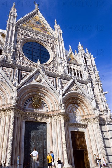 ITALY, Tuscany, Siena, The pink  black and white marble facade of the Duomo cathedral church of Santa Maria Assunta under a blue sky with tourists at the base by the doors.
