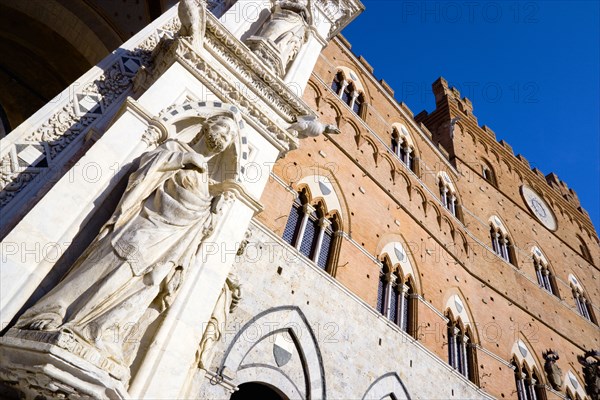 ITALY, Tuscany, Siena, Religious carvings on the portico entrance and facade of the Palazzo Publico town hall in the Piazza del Campo under a blue sky. The black and white crest or symbol of Siena decorates the facade of the building.