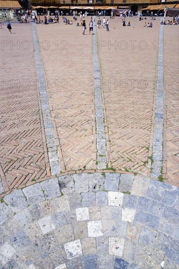ITALY, Tuscany, Siena, The Piazza del Campo and surrounding buildings with tourists walking in the square. Three lines in flat stone laid into the herringbone brick pattern of the square radiate from the central drain towards the buildings.