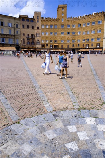 ITALY, Tuscany, Siena, The Piazza del Campo and surrounding buildings with tourists walking in the square. Three lines in flat stone laid into the herringbone brick pattern of the square radiate from the central drain towards the buildings.