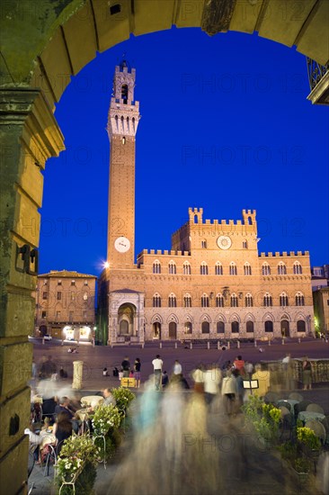 ITALY, Tuscany, Siena, The Torre del Mangia campanile belltower of the Palazzo Publico illuminated at night seen through the archway of the narrow sidestreet leading into the Piazza del Campo with people seated at tables and walking in the square.
