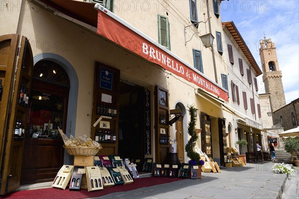 ITALY, Tuscany, Montalcino, Val D'Orcia Brunello di Montalcino Enoteca or wine shop with dispaly of boxed wines on the pavement around its entrance beneath a sunshade. The 14 the Century belltower of the Palazzo Comunale is at the end of the street.