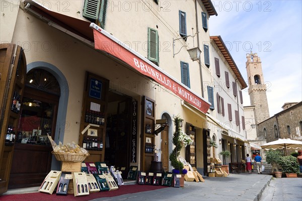 ITALY, Tuscany, Montalcino, Val D'Orcia Brunello di Montalcino Enoteca or wine shop with dispaly of boxed wines on the pavement around its entrance beneath a sunshade. The 14 the Century belltower of the Palazzo Comunale is at the end of the street.