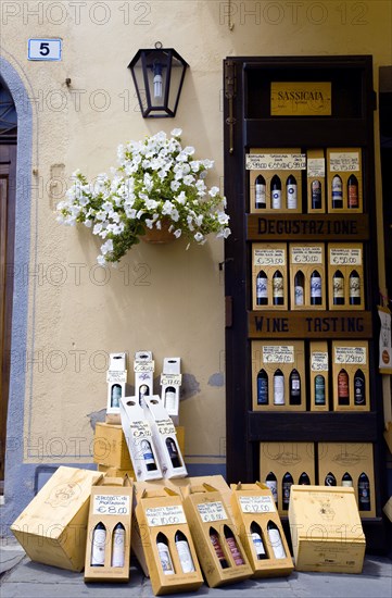 ITALY, Tuscany, Montalcino, Val D'Orcia Brunello di Montalcino Enoteca or wine shop with dispaly of boxed wines on the pavement by its entrance undera potted white gerranium plant beside a wine tasting sign.