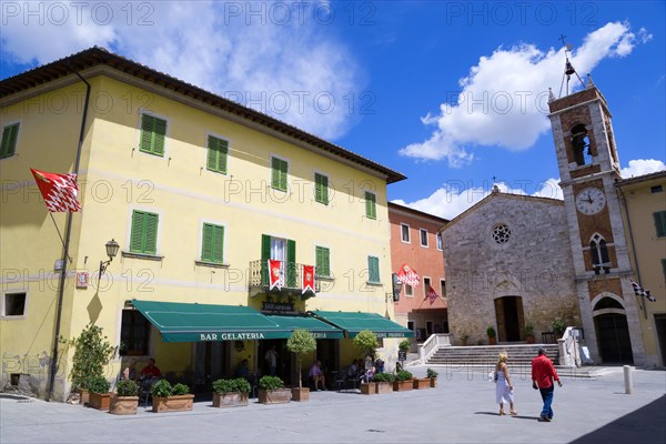 ITALY, Tuscany, San Quirico D'Orcia, Red and white flags of the Castello Quartieri or quarter decorate the Bar Centrale in the main square with a couple walking towards the Church of St Francis or Chiesa di San Francesco a San Quirico during the Festival of Barbarossa.