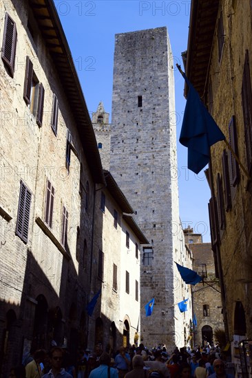 ITALY, Tuscany, San Gimignano, Via di Querececchio lined with flags and busy with tourists walking past shops towards two of the town's medieval towers.