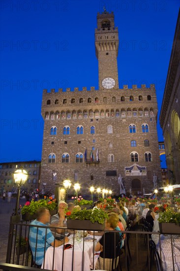 ITALY, Tuscany, Florence, People dining at restaurant tables outdoors at night in the Piazza della Signoria with the Campanile bell tower of the Palazzo Vecchio illuminated beyond.