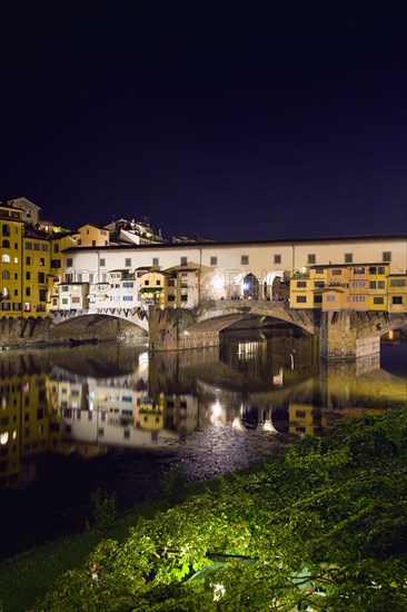 ITALY, Tuscany, Florence, Ponte Vecchio medieval bridge across River Arno illuminated at night with sightseeing tourists beside merchant's shops that line bridge and hang over the water below.