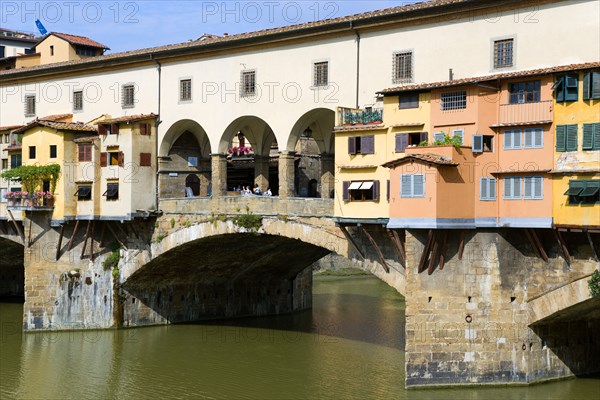 ITALY, Tuscany, Florence, Ponte Vecchio medieval bridge across River Arno with sightseeing tourists beside merchant's shops that line bridge and hang over the water below.