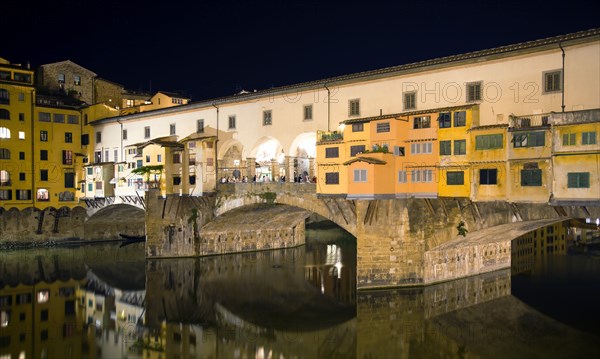ITALY, Tuscany, Florence, Ponte Vecchio medieval bridge across River Arno illuminated at night with sightseeing tourists beside merchant's shops that line bridge and hang over the water below.
