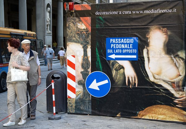 ITALY, Tuscany, Florence, Tourist couple walking past a billboard outside the Uffizi Gallery that conceals renovation work, the man staring open mouthed at the figure in the painting of a woman showing her naked breasts. Direction signs for pedestrians in foreground.