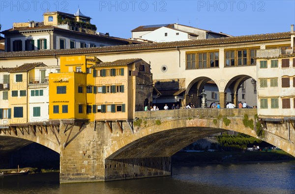 ITALY, Tuscany, Florence, Ponte Vecchio medieval bridge across River Arno with sightseeing tourists beside merchant's shops that line bridge and hang over the water below.