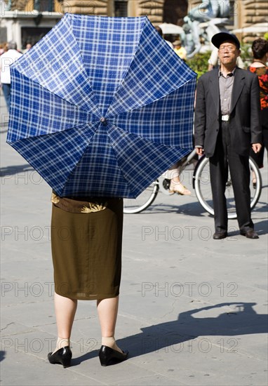 ITALY, Tuscany, Florence, Elderly senior Asian tourist couple in Piazza della Signoria with the woman under an umbrella for shade taking a photograph of the man.