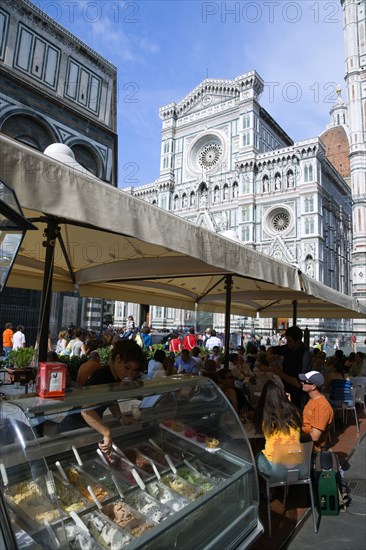 ITALY, Tuscany, Florence, The Neo-Gothic marble west facade of the Cathedral of Santa Maria del Fiore the Duomo with sightseeing tourists people sitting under umbrellas in the shade at a restaurant and a waitress serving ice cream from a counter.