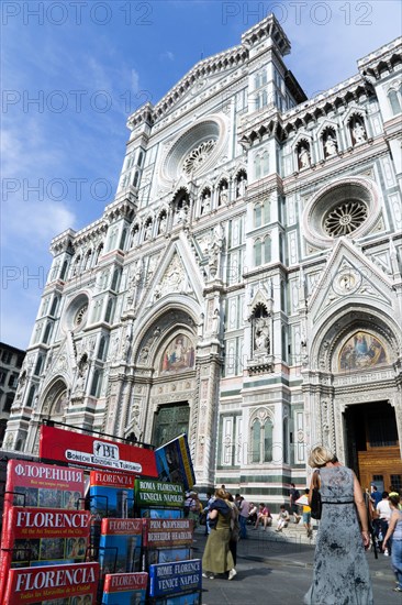 ITALY, Tuscany, Florence, The Neo-Gothic marble west facade of the Cathedral of Santa Maria del Fiore the Duomo with sightseeing tourists sitting on the steps and guidebooks in different languages at a souvenir stall in the foreground.
