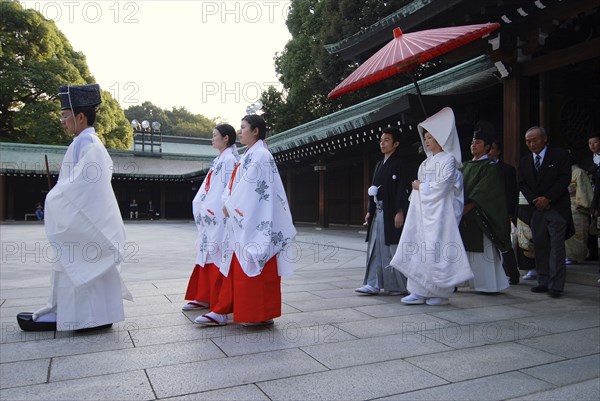Japan, Tokyo, Yoyogi, Meiji Jingu shrine, a wedding party led by a shinto priest and two woman attendants , bride in traditional white wedding kimono, groom in traditional kimono, both about thirty years old, shinto priest holds red umbrella over them,  familys follow.