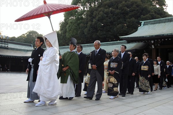 Japan, Tokyo, Yoyogi - at Meiji Jingu shrine, a wedding party in procession, bride in traditional white wedding kimono in front with groom in traditional kimono, about thirty years old, familys in rear, shinto priest holds red umbrella.