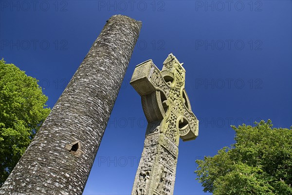 IRELAND, County Louth, Monasterboice Monastic Site, the West Cross slanted angular view of the east face with Round Tower in the background.