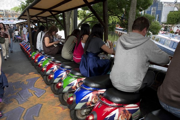 ENGLAND, London, Camden Town, People sitting on small colorful vespa scooters on a line formation under stalls, enjoying the view of Camden Towns canals, while eating their food and other people are walking behind them.