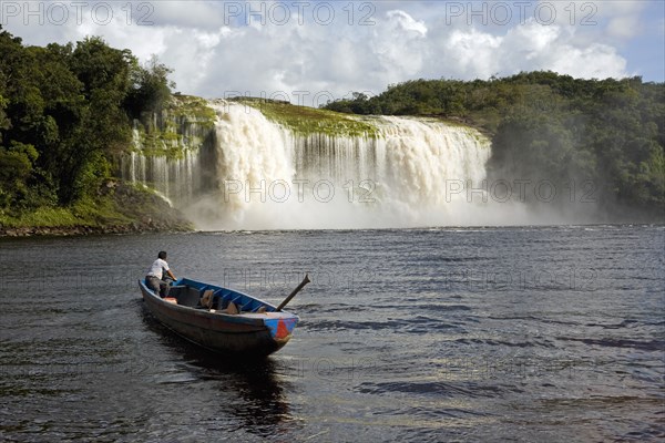 VENEZUELA, Bolivar State, Canaima National Park, Canaima Village, A Pemon tribe man while sailing with his boat at Canaima lake just in front of a big waterfall on a bright day with blue sky and white clouds.