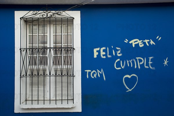 VENEZUELA, Bolivar State, Ciudad Bolivar, blue wall with graffiti words in Spanish and the symbol of a heart just next of a closed symmetrical window within a white painted frame, secured with iron bars.