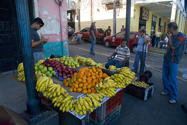 VENEZUELA, Bolivar State, Ciudad Bolivar, Fruit stall with bananas, oranges, apples and other fruits at Ciudad Bolivarís main street just by the Orinoco River with people looking at them and talking to the salesman while other people, cars and old buildings with columns are noticeable at the background. People using mobile phones.