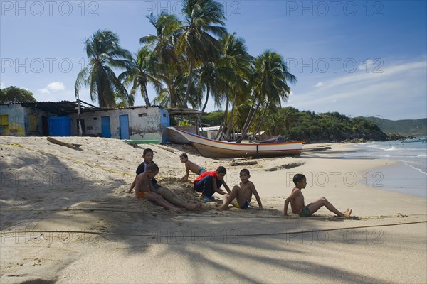 VENEZUELA, Margarita Island, Playa la Galera, Kids playing on the beach, under the shade of a palm tree just in front of the seawater while other trees, a house and a boat are noticeable at the background, shoot on a bright day with blue sky and some white clouds.
