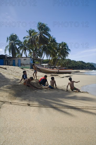 VENEZUELA, Margarita Island, Playa la Galera,  Kids playing on the beach, under the shade of a palm tree just in front of the seawater while other trees, a house and a boat are noticeable at the background, shoot on a bright day with blue sky and some white clouds.