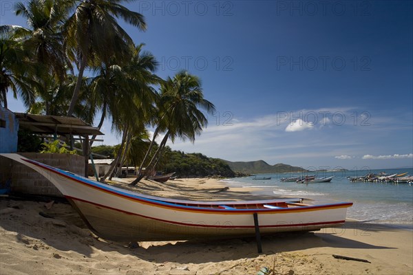 VENEZUELA, Margarita Island, Playa la Galera, Boat on the tropical beach just in front of a small house and palm trees while other boats are noticeable at the crystal clear seawater, shoot on a bright day with blue sky and white clouds.