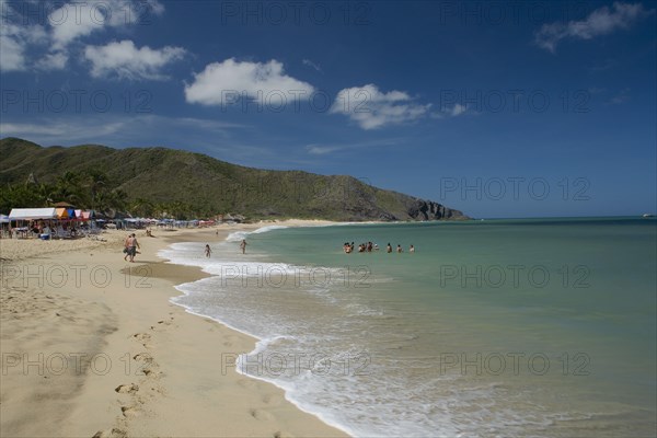 VENEZUELA, Margarita Island, Playa Caribe, View of exotic beach with people swimming and walking on the sand while deck chairs, bars and restaurants are noticeable, shoot on a bright day with blue sky and white clouds.