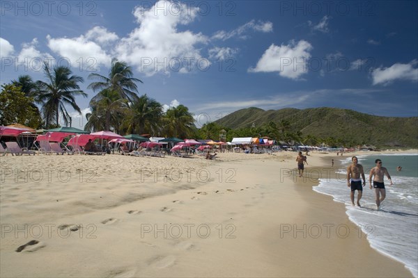 VENEZUELA, Margarita Island, Playa Caribe, View of exotic beach with people walking on the sand while colorful tents, deck chairs, bars, restaurants and palm trees are noticeable, on a bright day with blue sky and white clouds.