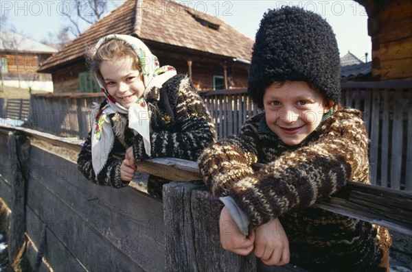 Romania, Maramuresh, Portrait of two local children wearing traditional clothing leaning over wooden fence.